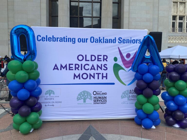 A banner for Older Americans Month and Aging Unbound is displayed outside, decorated with blue balloon columns shaped like “O” and “A,” plus logos for the City of Oakland, Human Services Department, and Mercy Brown Bag.