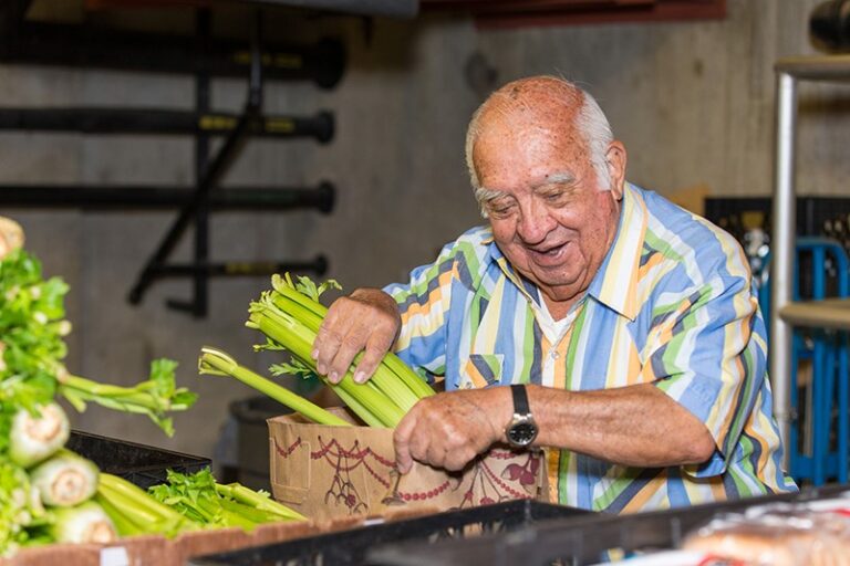 An elderly man places fresh celery stalks into a box in what appears to be a produce storage or market area, highlighting the idea of using food for health.