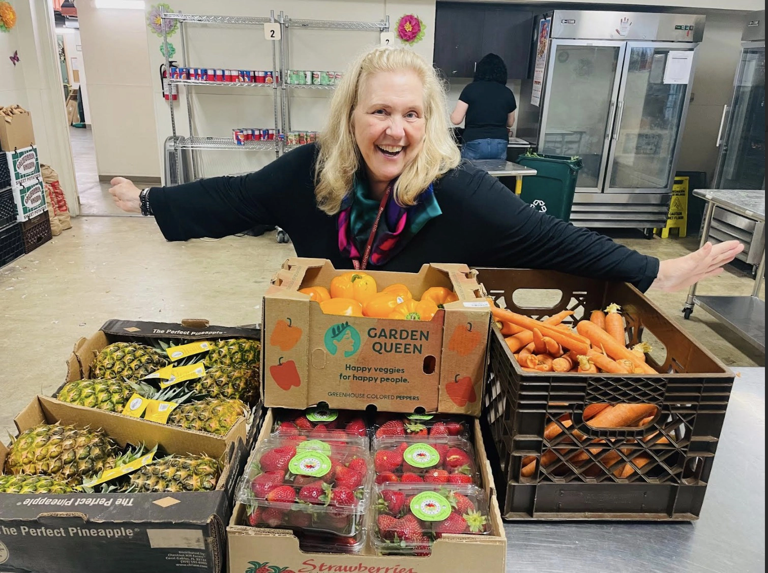 A woman stands behind crates of fresh produce—pineapples, strawberries, oranges, and carrots—in a food distribution area, highlighting the Food as Medicine approach to health and wellness.