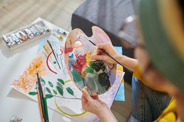 Person mixing paint on a palette next to an abstract watercolor painting, embodying how art can inspire creativity and support healthy aging, with tubes of paint and brushes on a white table.