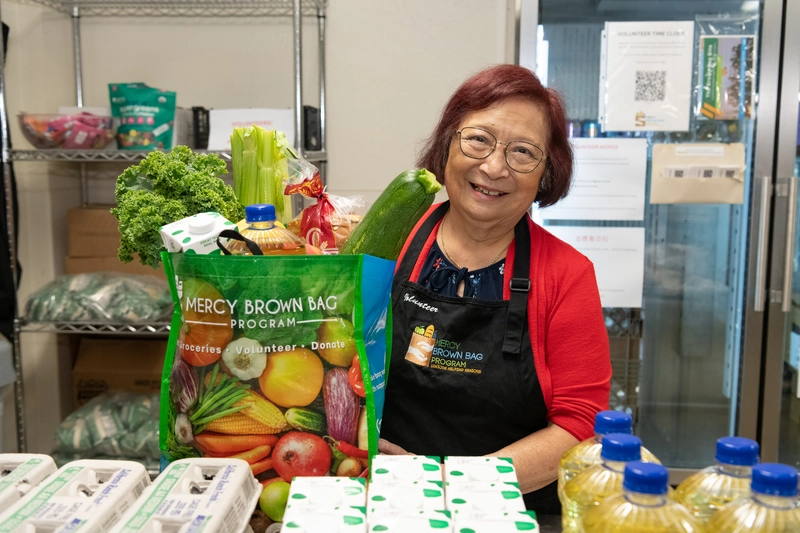 A woman wearing a Mercy Brown Bag Program apron stands in a food pantry, smiling beside a reusable bag filled with fresh produce and groceries.