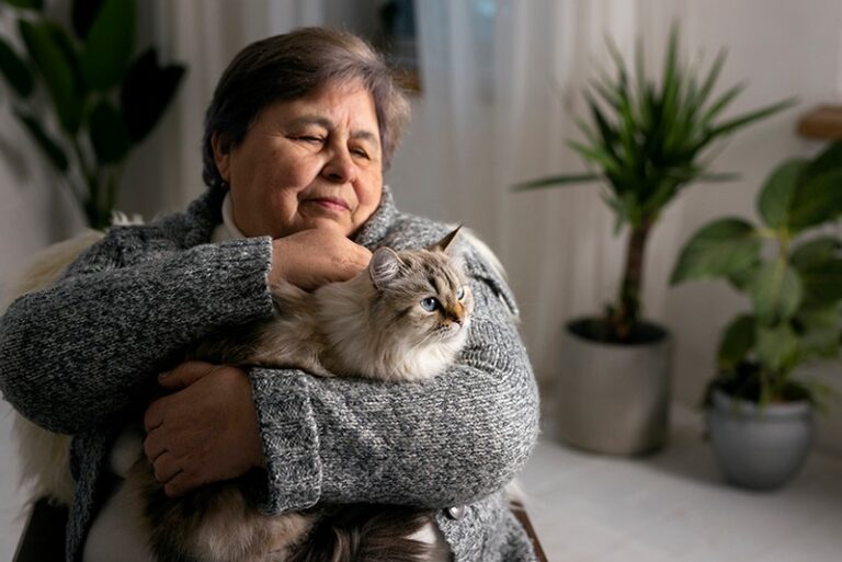 An older woman in a gray sweater holds her fluffy cat, enjoying the companionship of pets while sitting indoors, with green potted plants in the background.