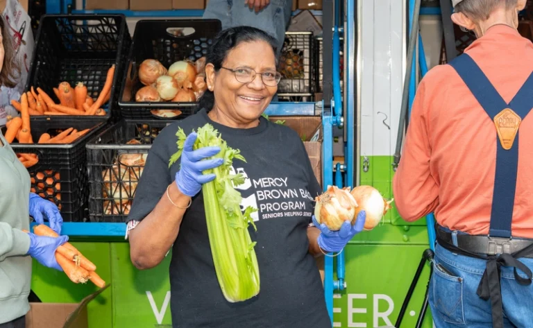 Woman in gloves holds celery and onions in front of crates of vegetables at a food distribution event, smiling at the camera.