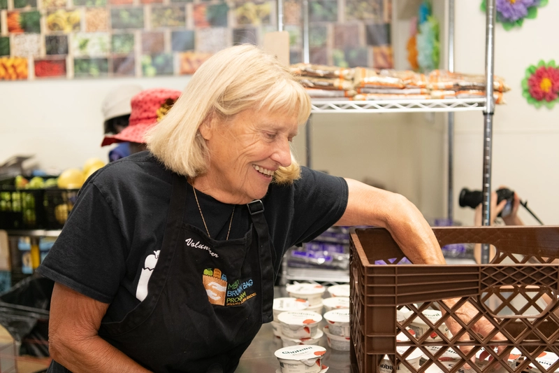 An older woman wearing a black apron smiles as she arranges food containers on a shelf in a pantry or food distribution setting.