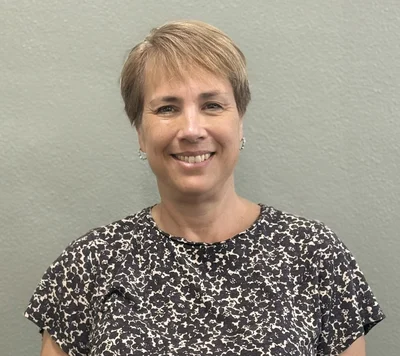 A woman with short, light brown hair is smiling at the camera. She is wearing a black and white patterned shirt and standing in front of a plain, light gray wall.