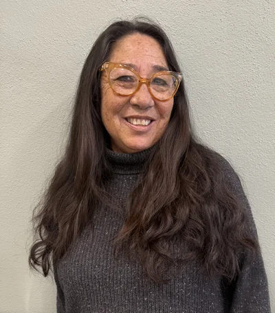 A woman with long brown hair and glasses smiles while standing against a plain, light-colored wall. She is wearing a dark gray turtleneck sweater.