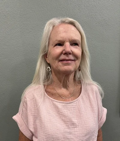 An older woman with long white hair is standing in front of a gray textured wall. She is wearing a light pink short-sleeve top, earrings, and a thin necklace, and she is looking slightly off camera with a slight smile.
