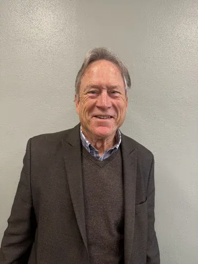 An older man with gray hair is standing in front of a plain light-colored wall, wearing a dark blazer over a brown sweater and a checkered shirt, and smiling at the camera.