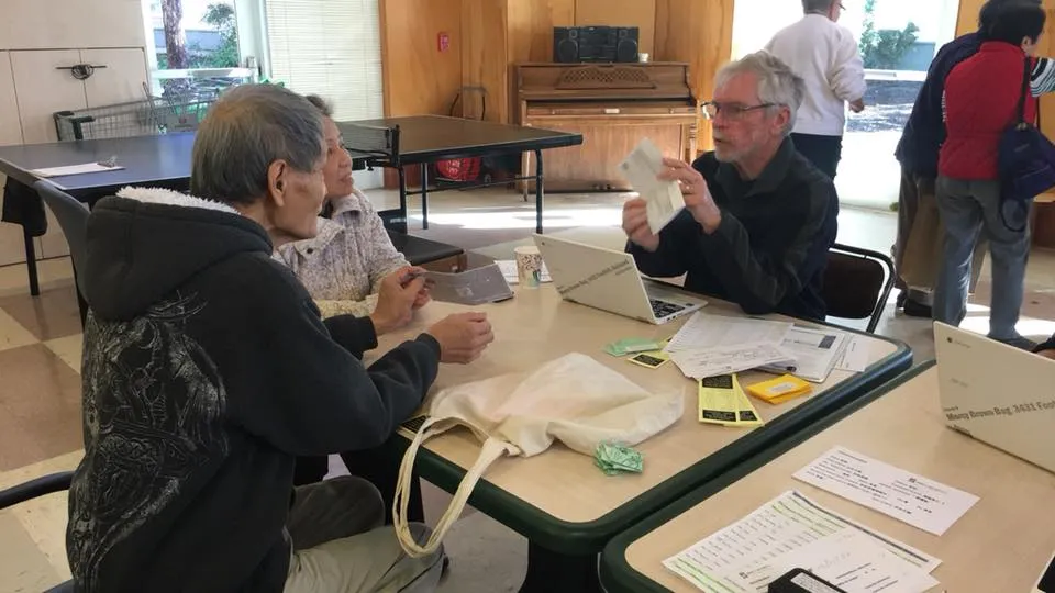 Three people sit at a table covered with papers and forms, engaged in discussion about the reality of injustice inside a community center with a ping pong table and piano in the background.