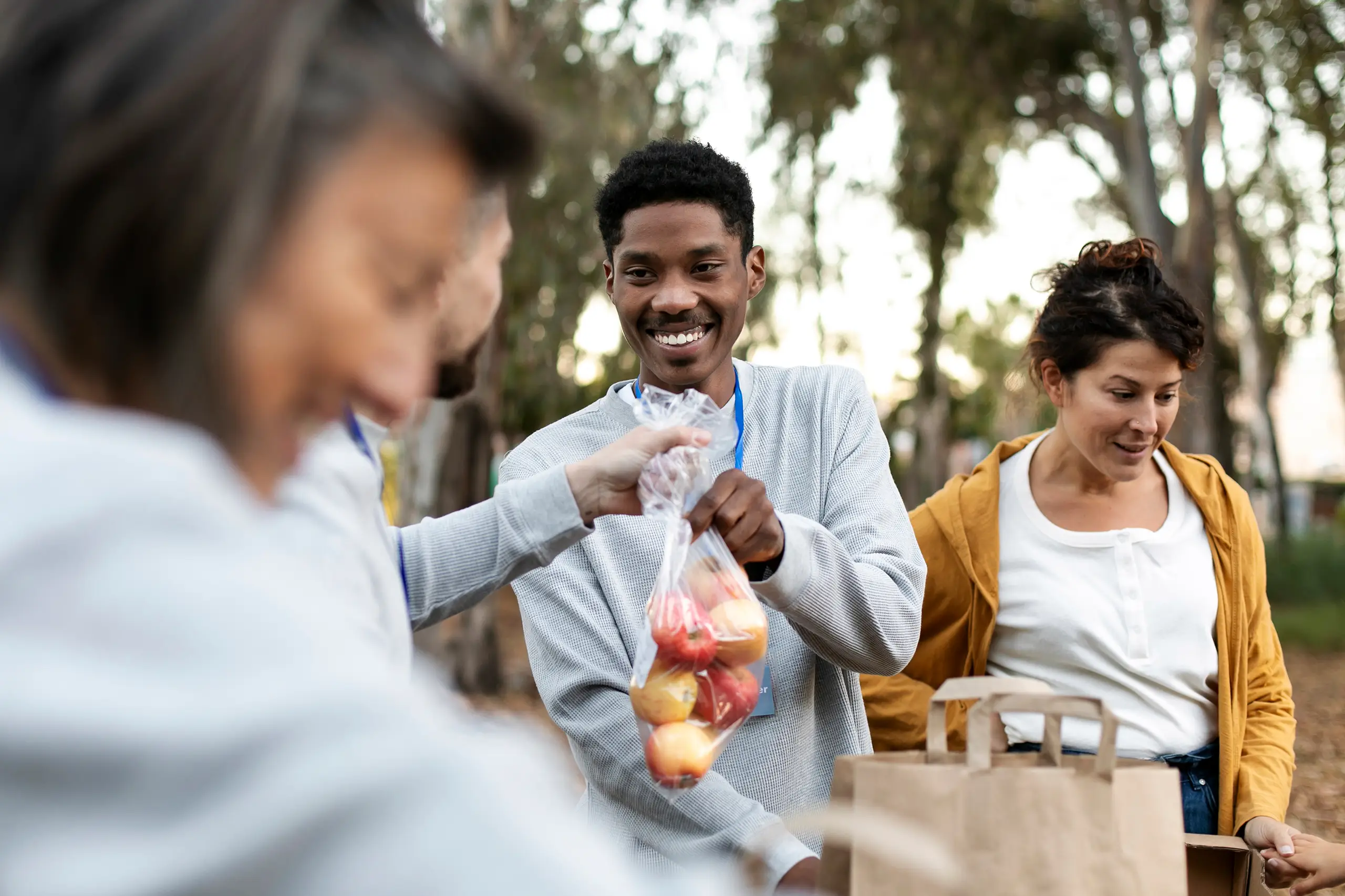 A man smiles while receiving a bag of apples from a volunteer at an outdoor food distribution event, with others and paper bags visible nearby.