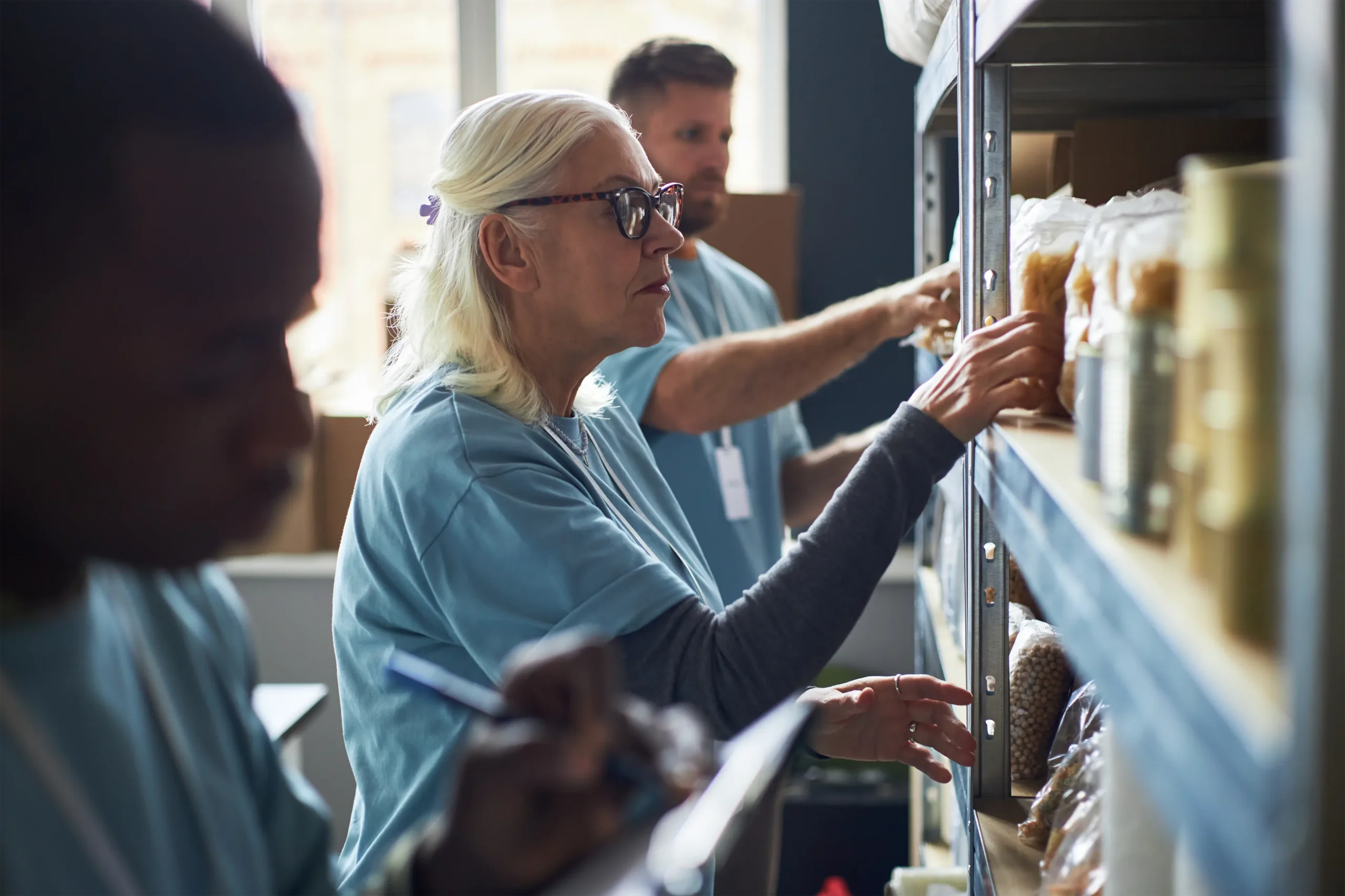 Three people in matching shirts organize and take inventory of food items on shelves in a storage room, showing how easy it is to Get Involved and make a difference.