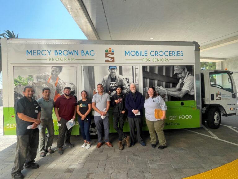 A group of people stands in front of Mercy Brown Bag Program’s new mobile groceries truck for seniors, parked under a concrete overhang.